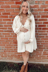 woman wearing a cream maternity dress with lace details and buttons, holding her baby bump against a brick wall