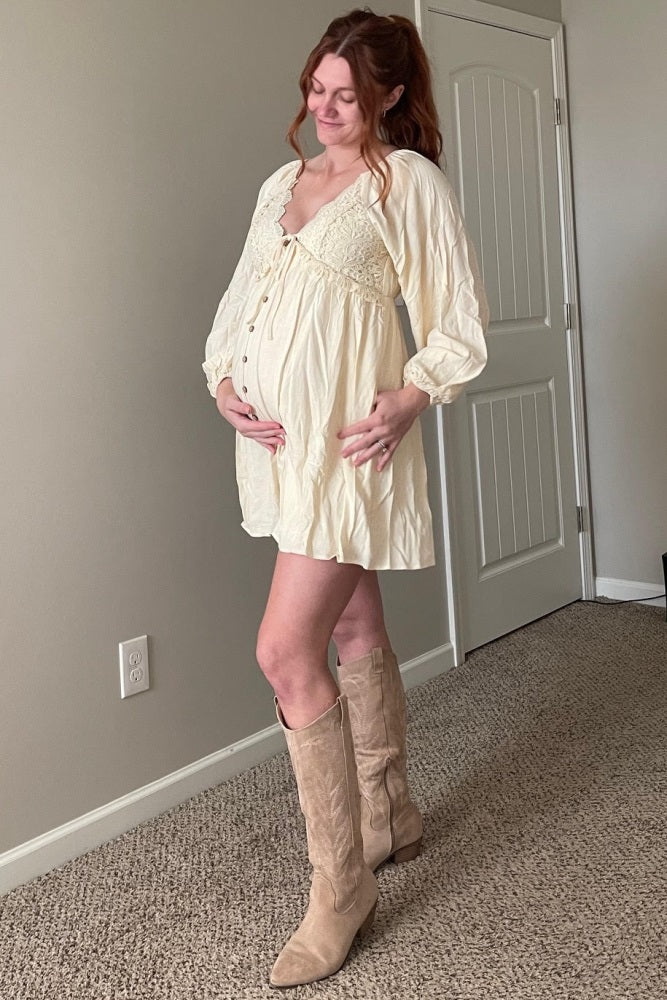 woman wearing a cream maternity dress with lace details, showing a baby bump, paired with knee-high beige boots