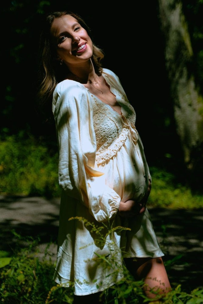 woman wearing a light yellow maternity dress with lace details and holding her baby bump in a forest setting