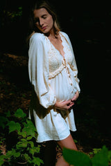 woman wearing a cream maternity dress with lace detail and buttons, cradling her baby bump in a natural setting