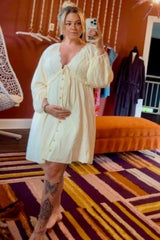 woman wearing a cream maternity dress with button details and a baby bump, posing in front of a mirror