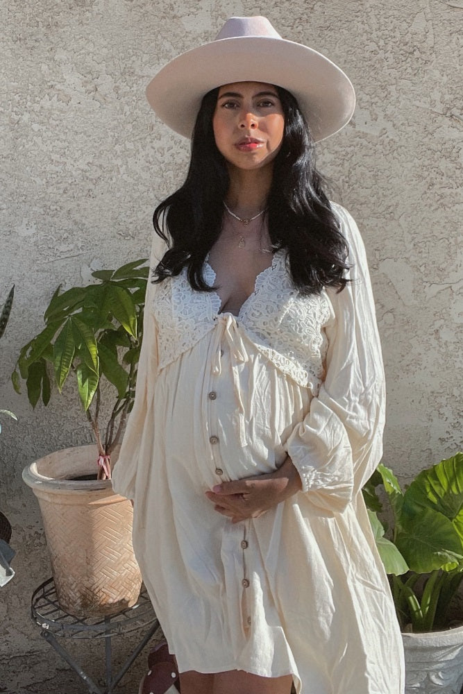 woman wearing a cream maternity dress with lace details and buttons, holding her baby bump while wearing a wide-brimmed hat