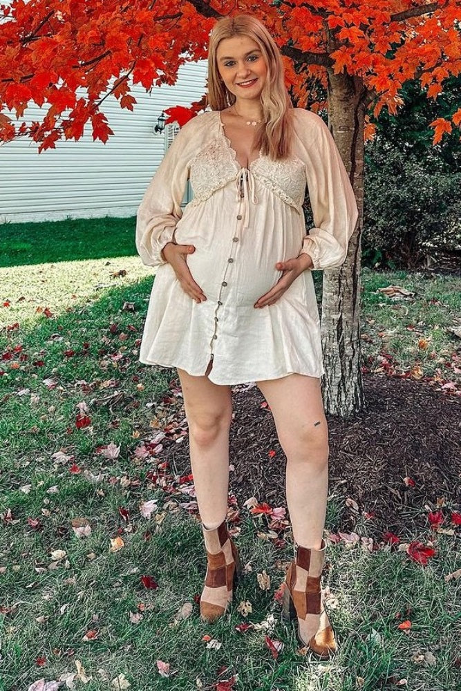 woman wearing a light beige maternity dress with a tie front and buttons, holding her baby bump outdoors