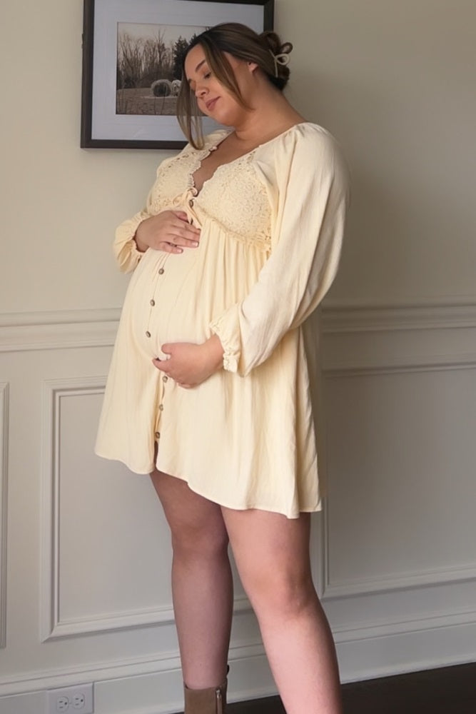 pregnant woman in a yellow lace maternity dress holding her baby bump in front of a white wall