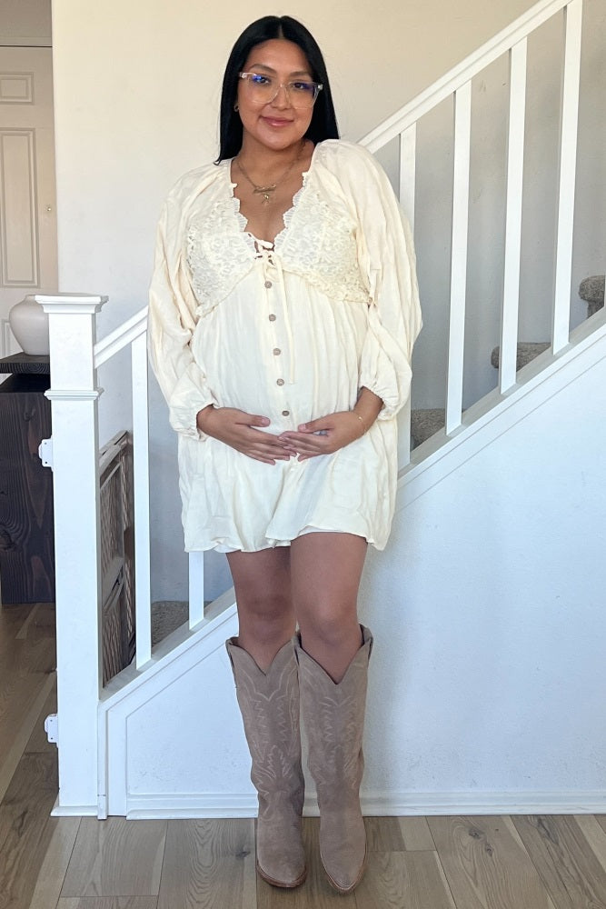 woman wearing a cream maternity dress with lace details, holding her baby bump, standing near a staircase