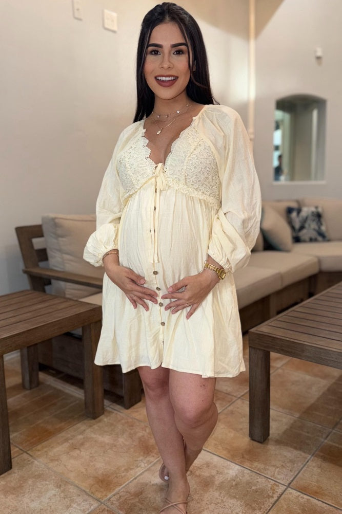 Smiling pregnant woman in a cream lace maternity dress holding her baby bump in a living room setting