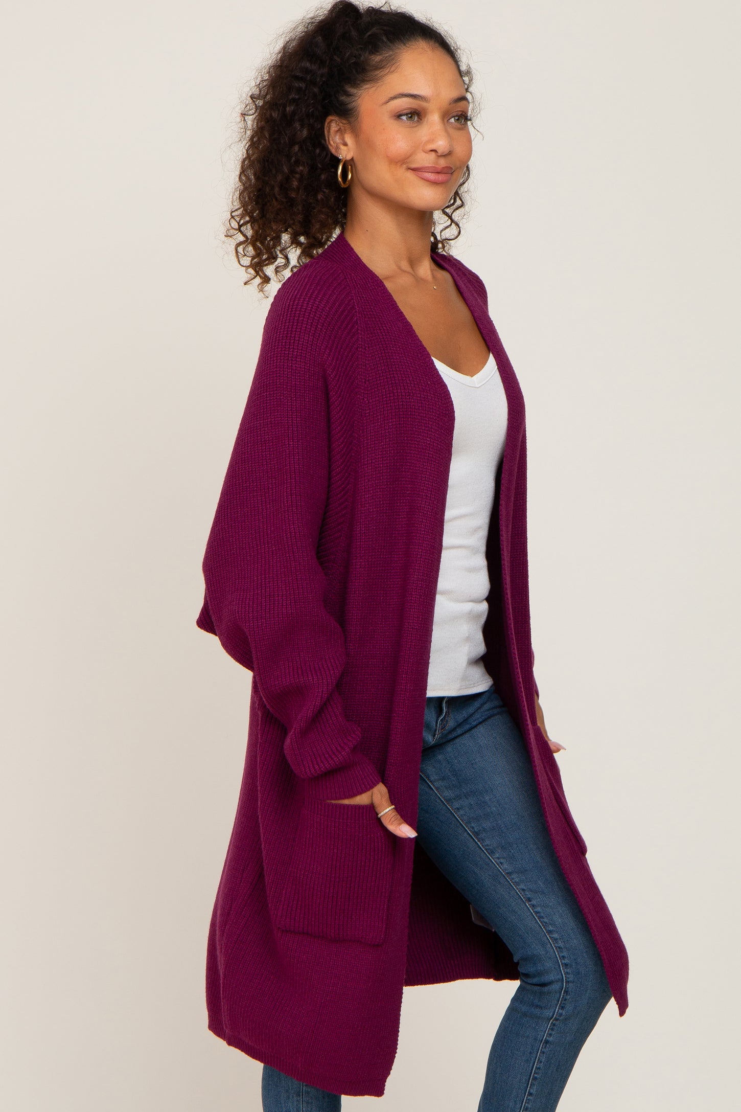 woman wearing a long purple cardigan over a white top and blue jeans, standing sideways with a relaxed expression