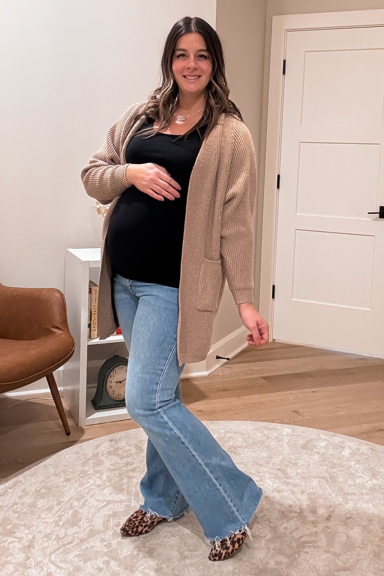 woman wearing a black tank top and beige cardigan, showcasing her baby bump with flared blue jeans