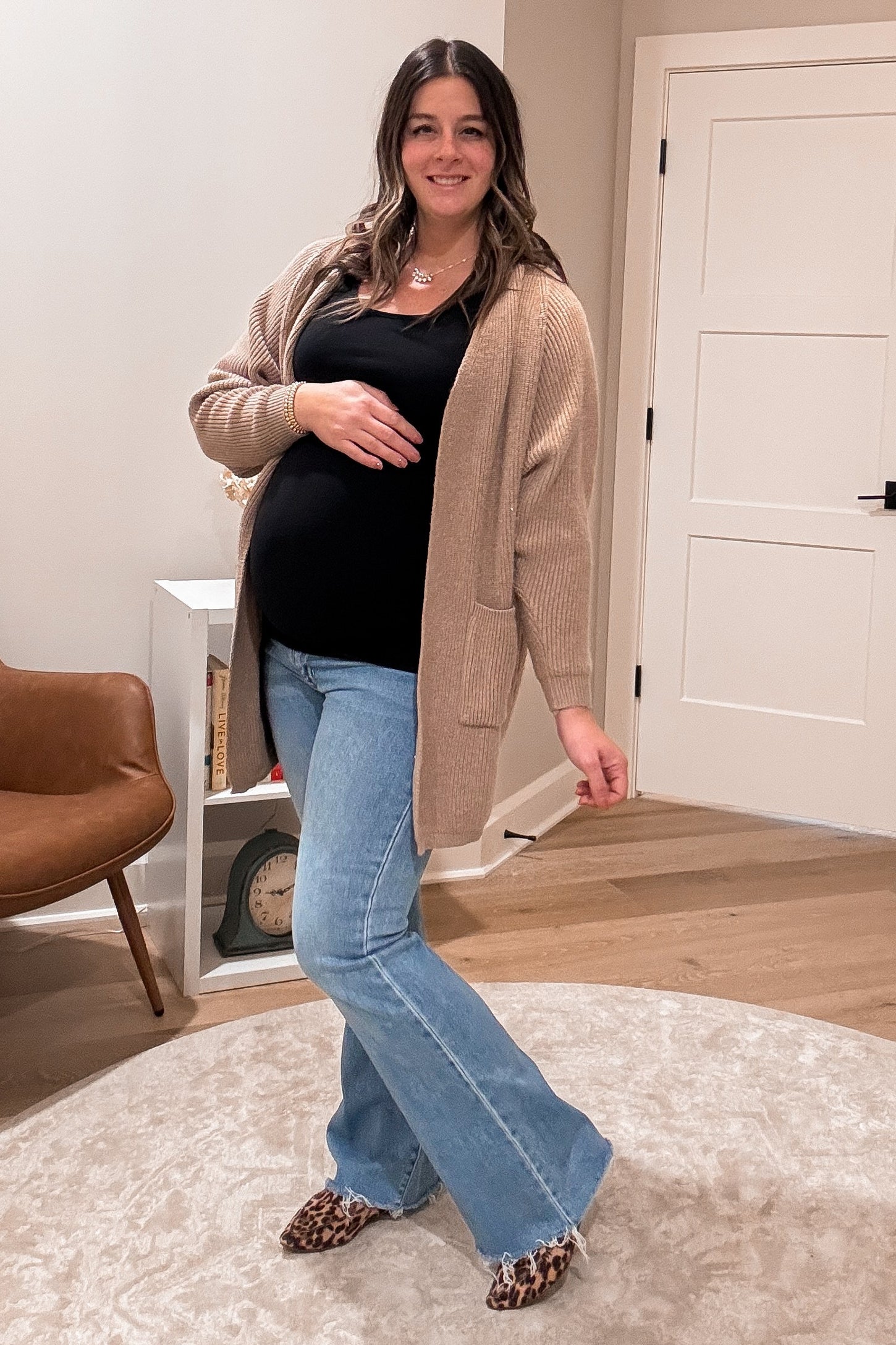 woman wearing a black tank top and beige cardigan, showcasing her baby bump with flared blue jeans