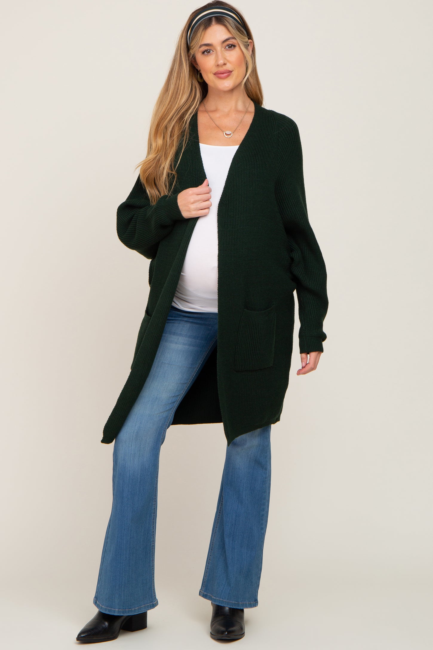 woman wearing a dark green maternity cardigan over a white top, with a visible baby bump, paired with blue flared jeans