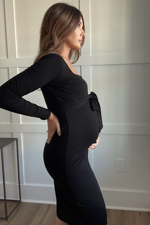 Pregnant woman in a long-sleeved black maternity dress with a side bow, posing against a white paneled wall.
