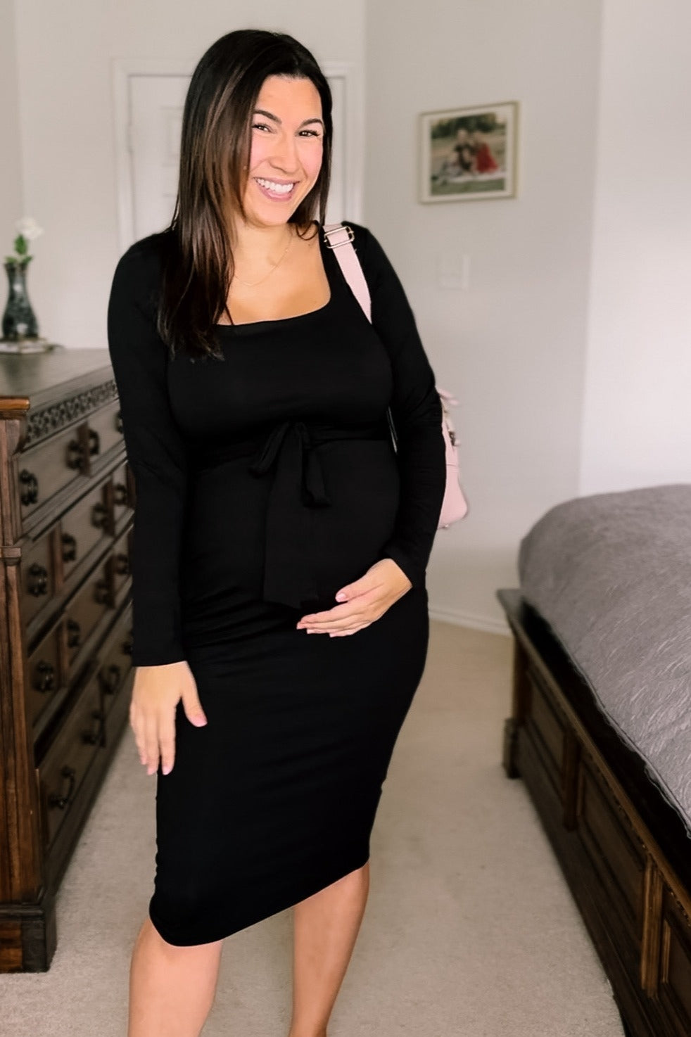 woman wearing a black long sleeve maternity dress with a tie waist and baby bump visible, smiling and holding her belly