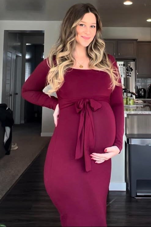 woman wearing a burgundy long sleeve maternity dress with a tie waist and holding her baby bump