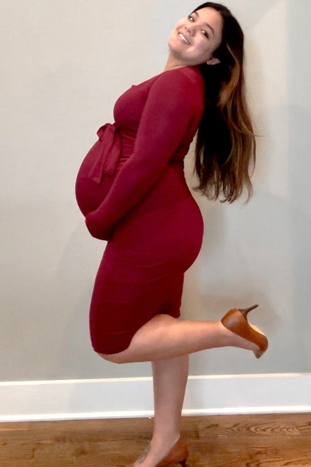 woman wearing a burgundy maternity bodycon dress with a tie waist and holding her baby bump
