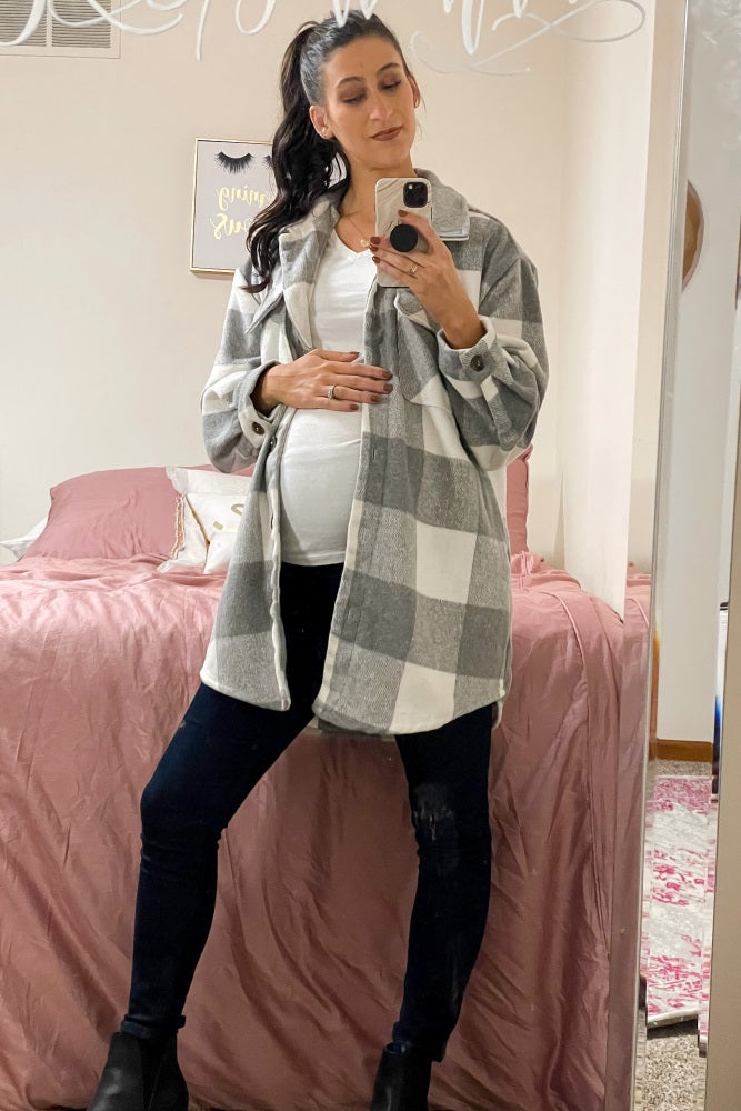 Pregnant woman in a grey and white plaid shacket, white t-shirt, and black jeans taking a mirror selfie in a bedroom