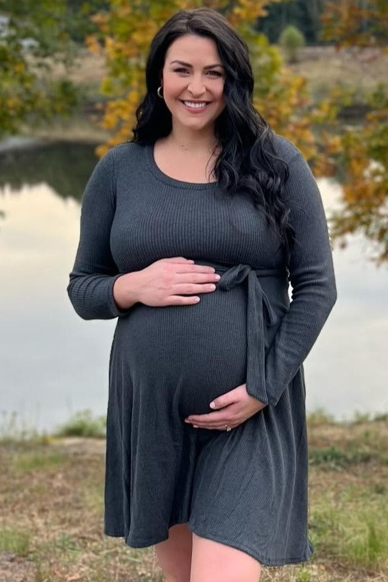 woman wearing a dark gray maternity dress with tie detail, holding her baby bump outdoors by a body of water