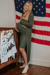 woman wearing an olive green maternity dress with long sleeves, holding her baby bump next to a crib and an American flag