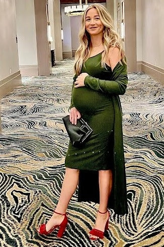 woman wearing a dark green maternity dress with an open cardigan, holding a clutch, and showing her baby bump