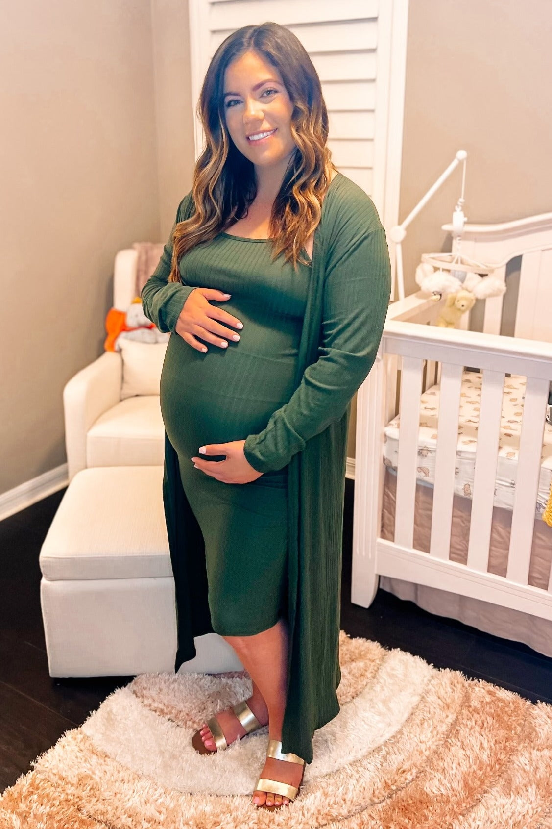 woman wearing a dark green maternity dress with a matching long cardigan and holding her baby bump in a nursery setting