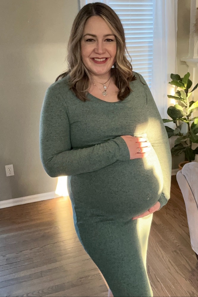 Smiling pregnant woman in a green long-sleeve maternity dress holding her baby bump in a living room