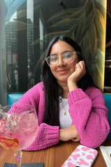 woman wearing a white top and a pink knit cardigan, sitting at a table with a cocktail and phone, smiling