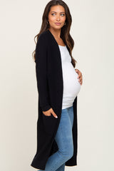 woman wearing a black long cardigan over a white maternity top, cradling her baby bump, paired with light blue jeans