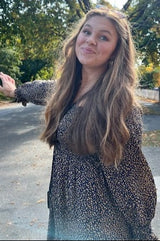 woman wearing a black floral long sleeve dress with a belted waist, standing outdoors with trees in the background