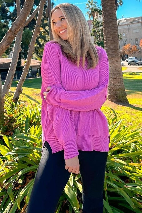 woman wearing a bright pink oversized sweater and black leggings, smiling in a park surrounded by greenery