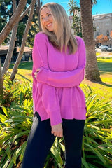woman wearing a bright pink oversized sweater and black leggings, smiling in a park surrounded by greenery