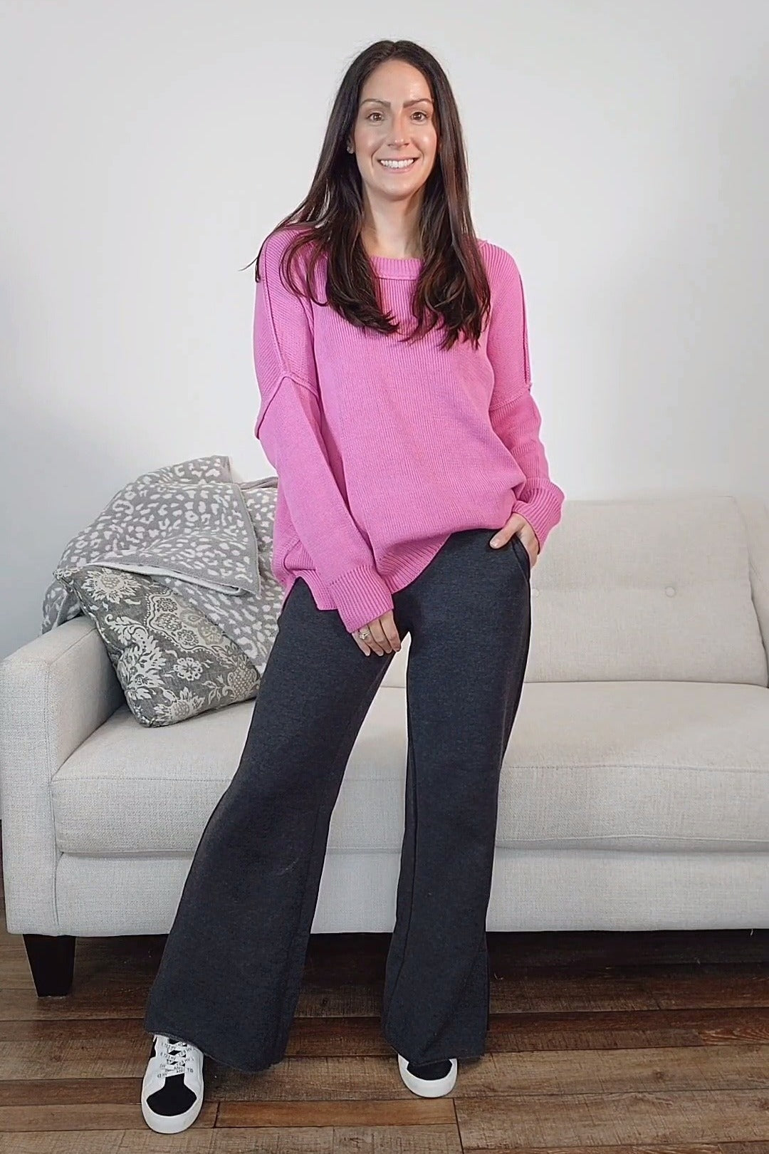 woman wearing a pink long sleeve sweater and gray wide-leg pants, standing in front of a sofa