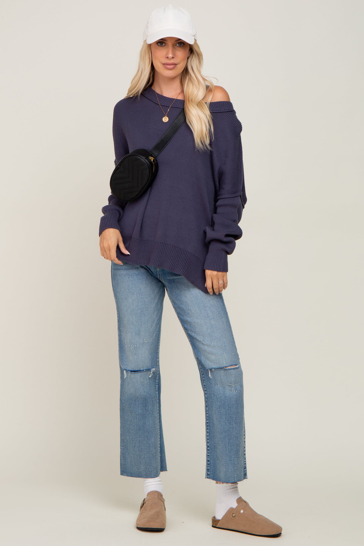 woman wearing a dark purple off-the-shoulder sweater, light blue cropped jeans, and a white cap with a black crossbody bag