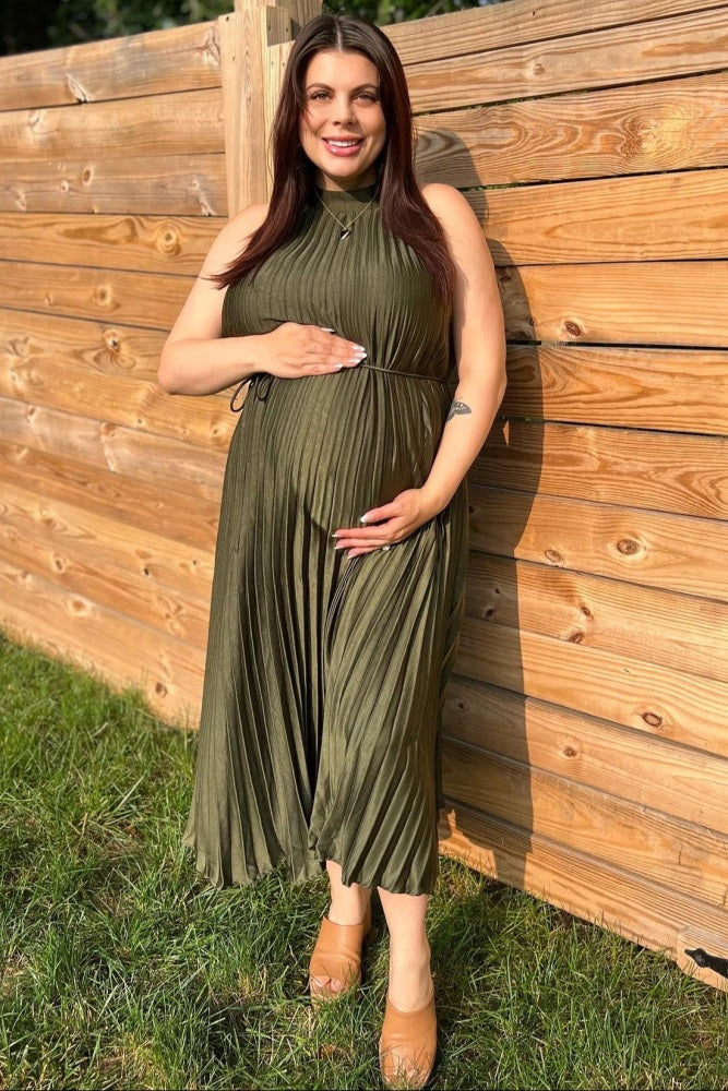 woman wearing an olive green pleated maternity dress, holding her baby bump, standing in grass near a wooden fence