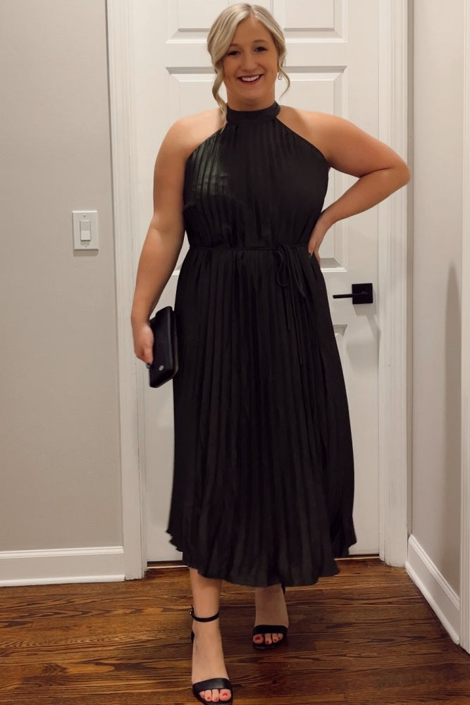 woman wearing a dark green pleated halter neck maternity dress, smiling and holding a black clutch