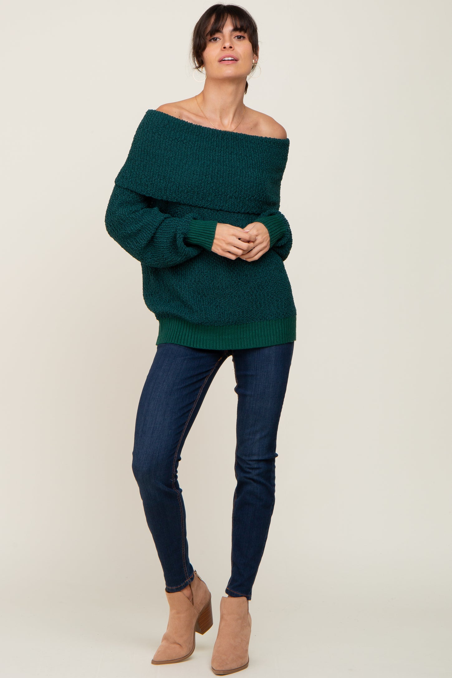 Woman wearing a dark green off-the-shoulder knit sweater, dark blue skinny jeans, and tan suede ankle boots.