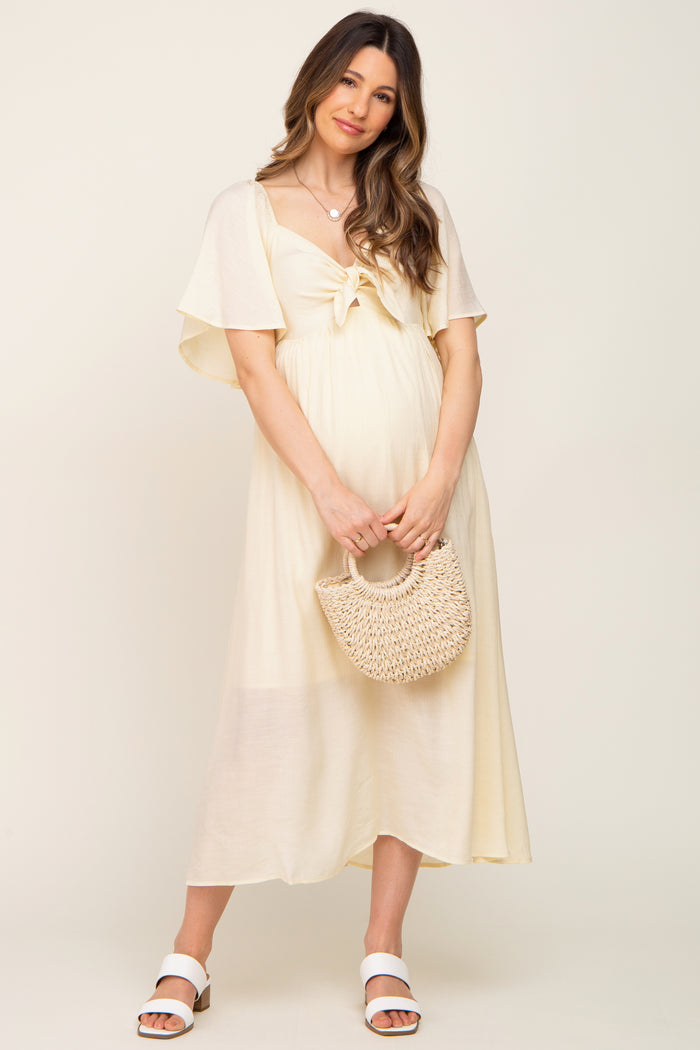 woman wearing a light yellow maternity dress with flutter sleeves and a knot detail, holding a woven handbag and showing her baby bump