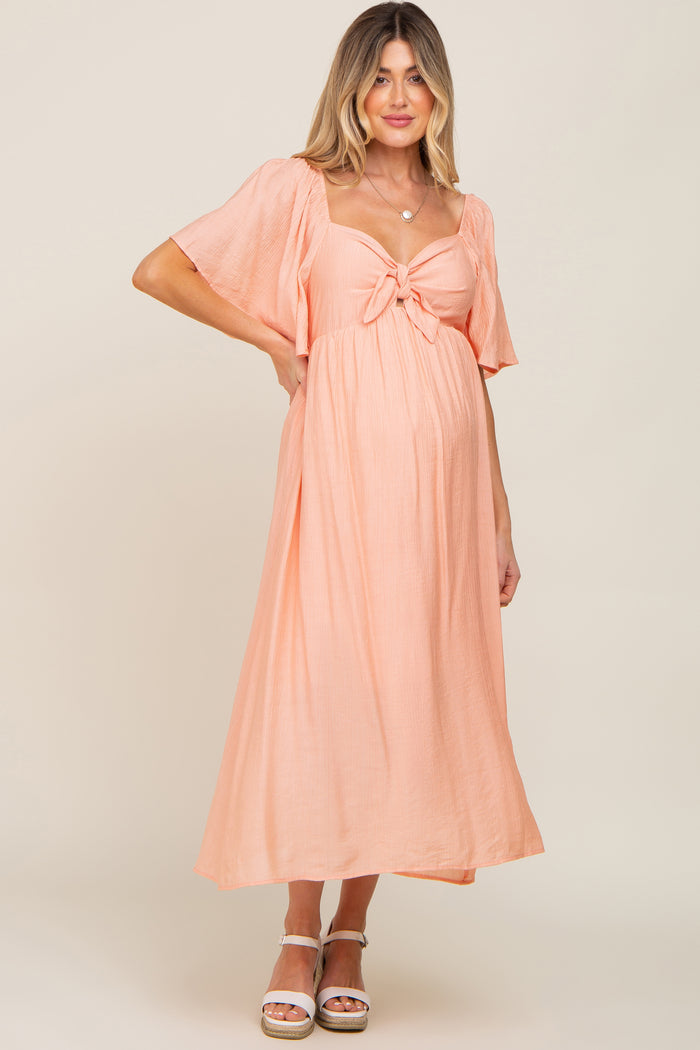 woman wearing a peach maternity maxi dress with a tie detail and flutter sleeves, showing her baby bump