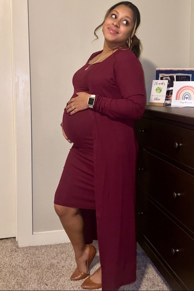 woman wearing a burgundy maternity dress with a long cardigan, holding her baby bump and smiling