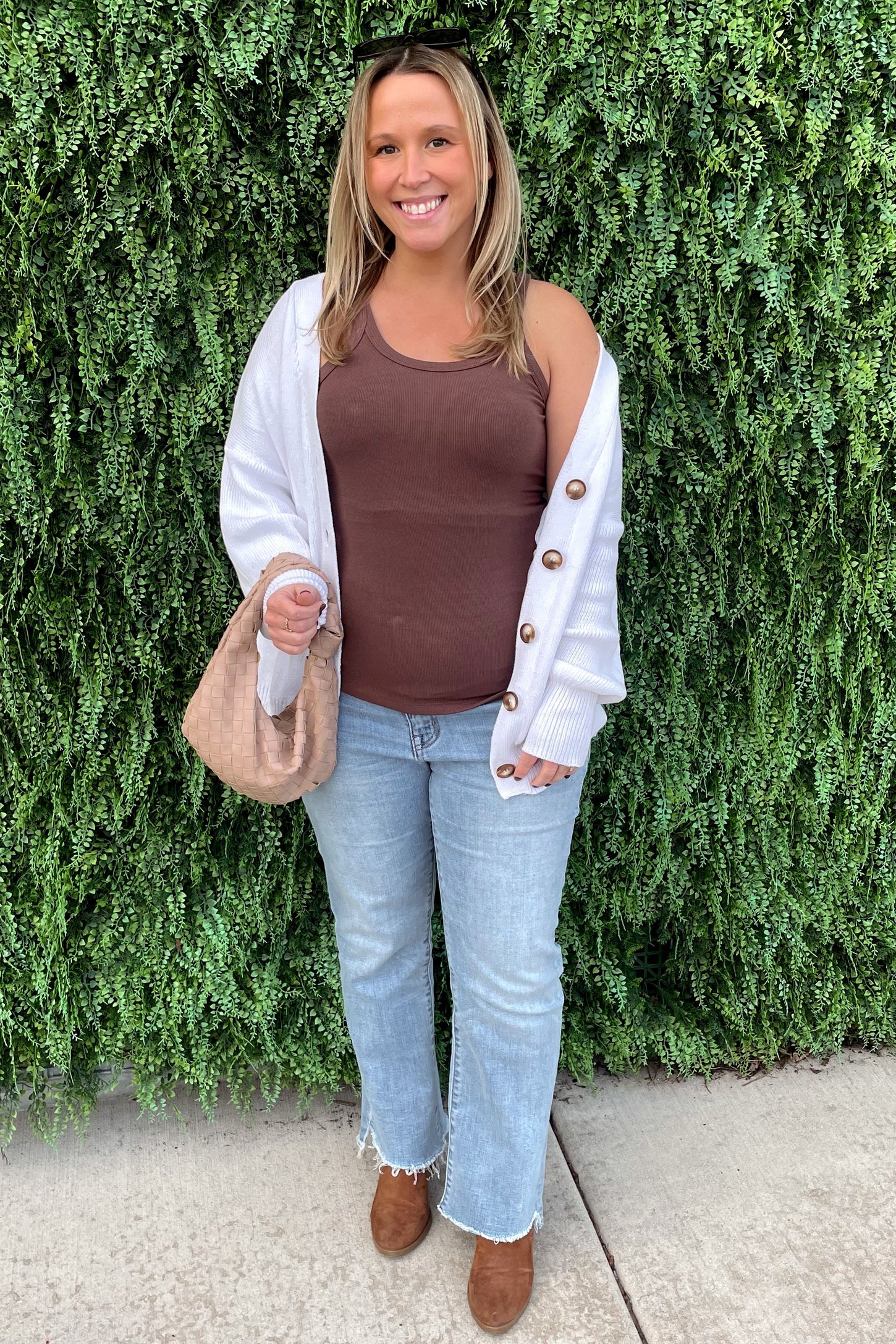 woman wearing a brown ribbed tank top, white cardigan, and light blue flared jeans, holding a tan bag, standing against greenery