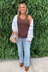woman wearing a brown ribbed tank top, white cardigan, and light blue flared jeans, holding a tan bag, standing against greenery