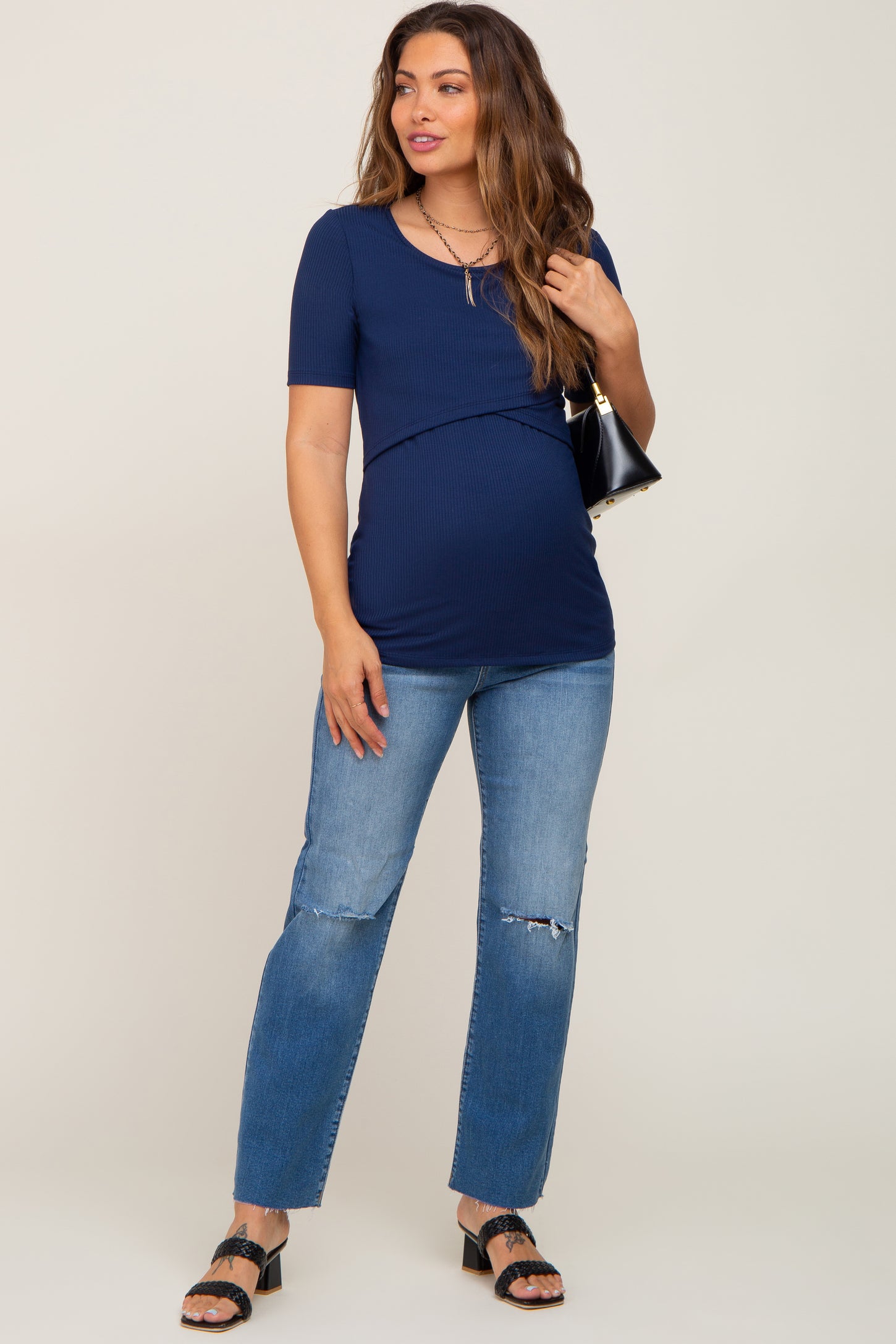 woman wearing a navy blue short sleeve maternity top with a baby bump and distressed jeans, holding a black handbag