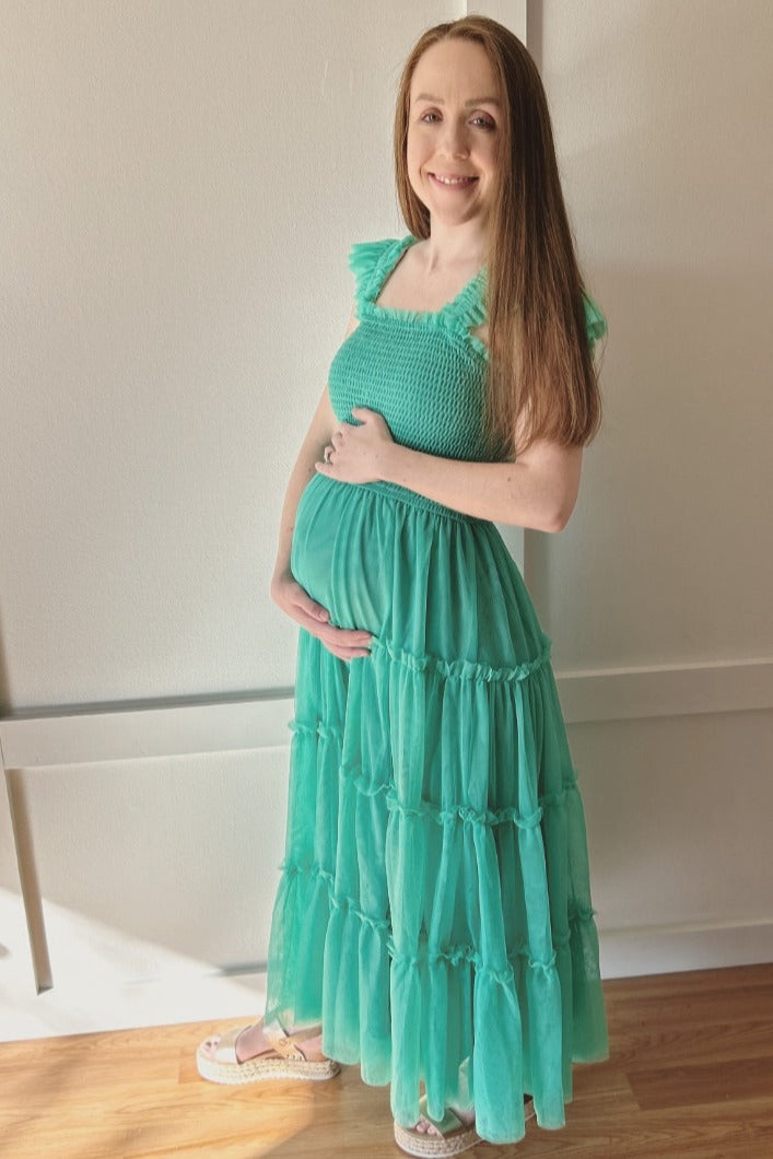 woman wearing a green tiered maternity dress with a smocked bodice and holding her baby bump