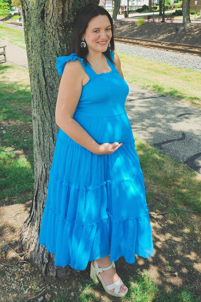 woman wearing a bright blue maternity dress with ruffled layers, standing by a tree and holding her baby bump