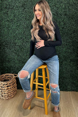woman wearing a black long sleeve maternity top and distressed jeans, sitting on a stool with visible baby bump