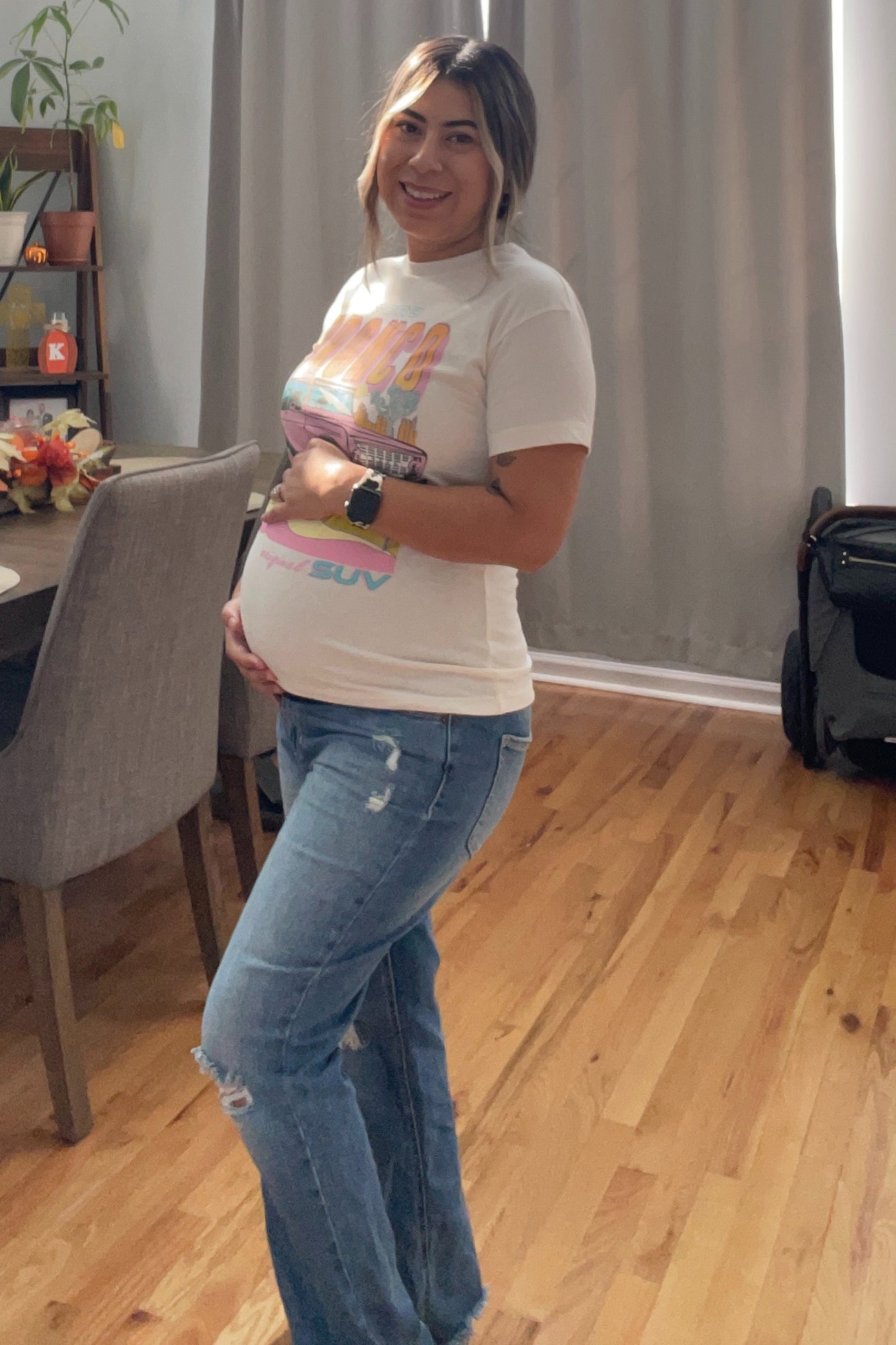 woman wearing a white graphic maternity t-shirt and blue flared jeans, showing her baby bump while standing in a modern kitchen