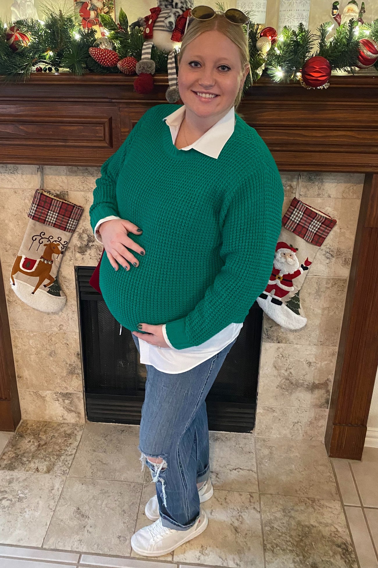 woman wearing a green chunky knit maternity sweater and white shirt, holding her baby bump in front of a festive mantle