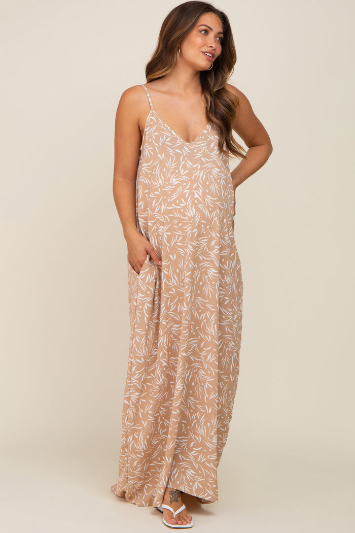 woman wearing a tan floral maternity maxi dress with adjustable straps and side pockets, showcasing her baby bump