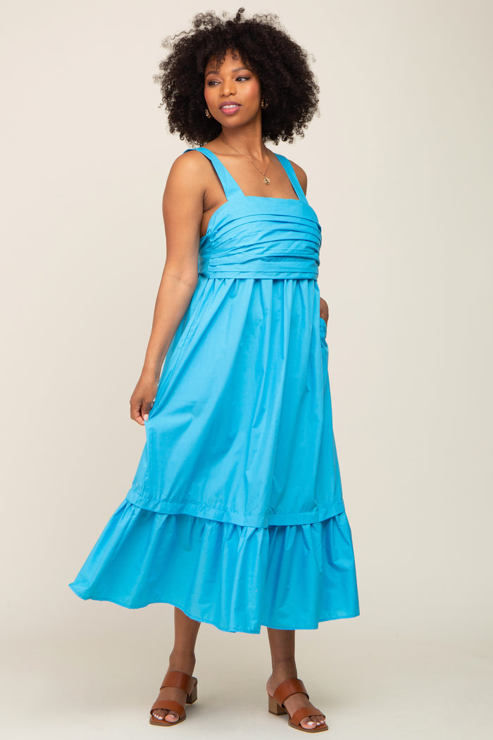 Woman with curly hair wearing a bright blue sleeveless midi dress with a pleated bodice and tiered skirt