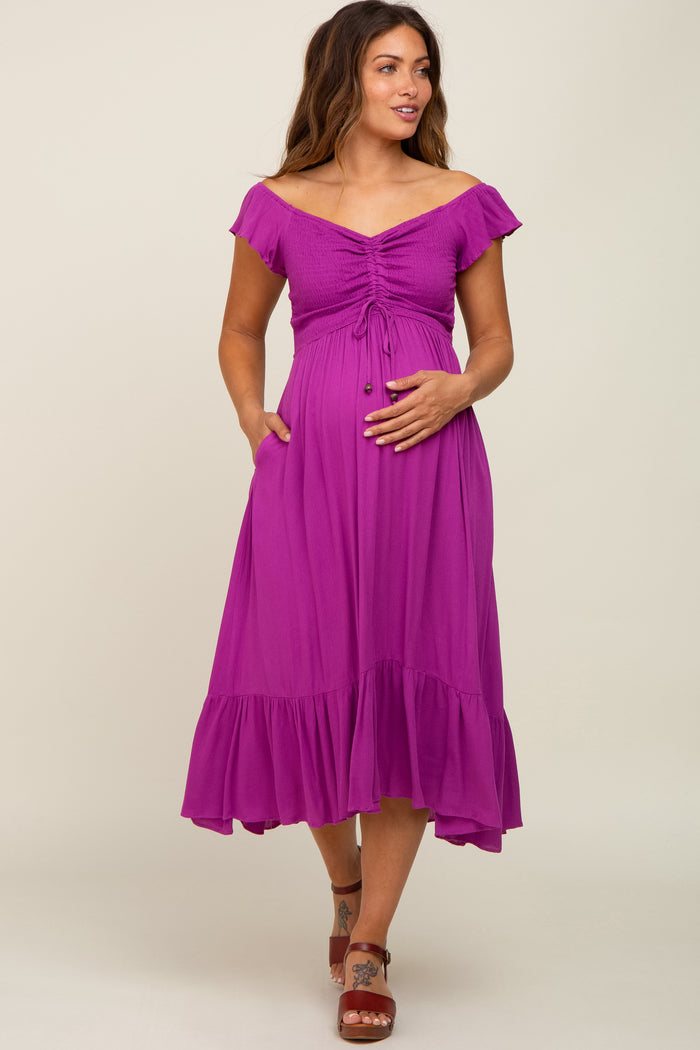 woman wearing a fuchsia off-the-shoulder maternity dress with a tie-front detail and ruffled hem, holding her baby bump