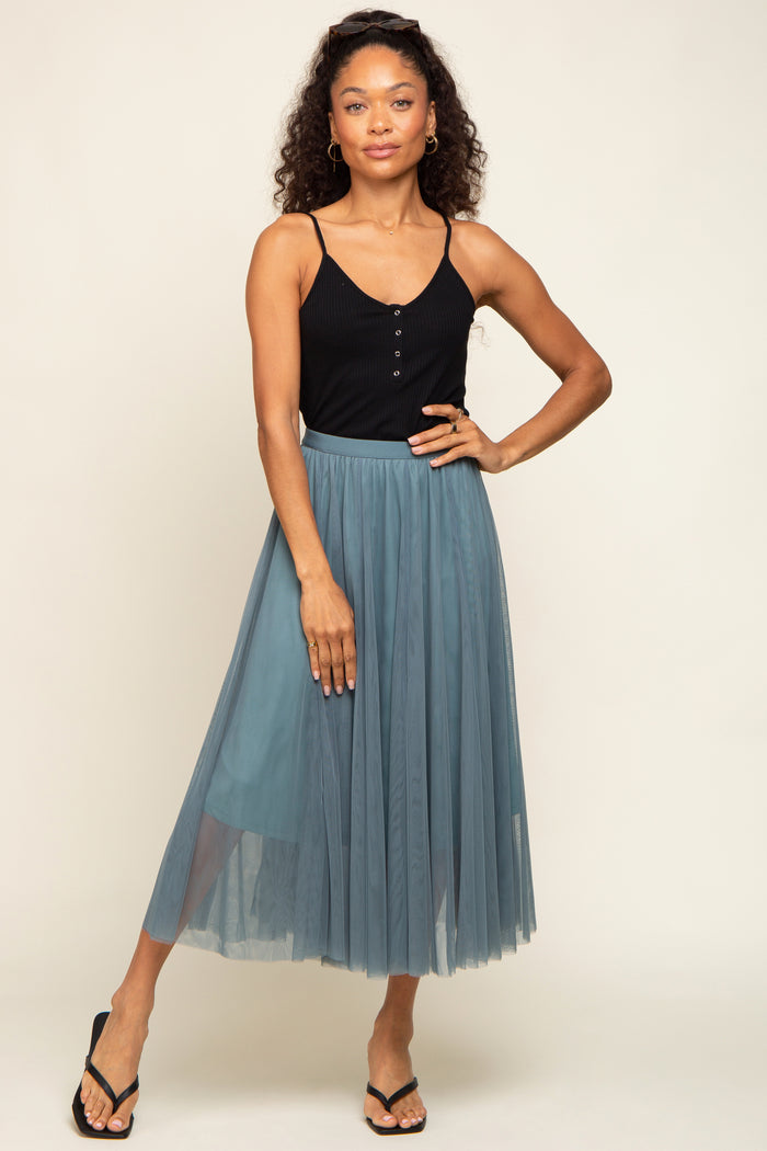 woman wearing a black fitted tank top and a teal tulle midi skirt, posing with one hand on her hip