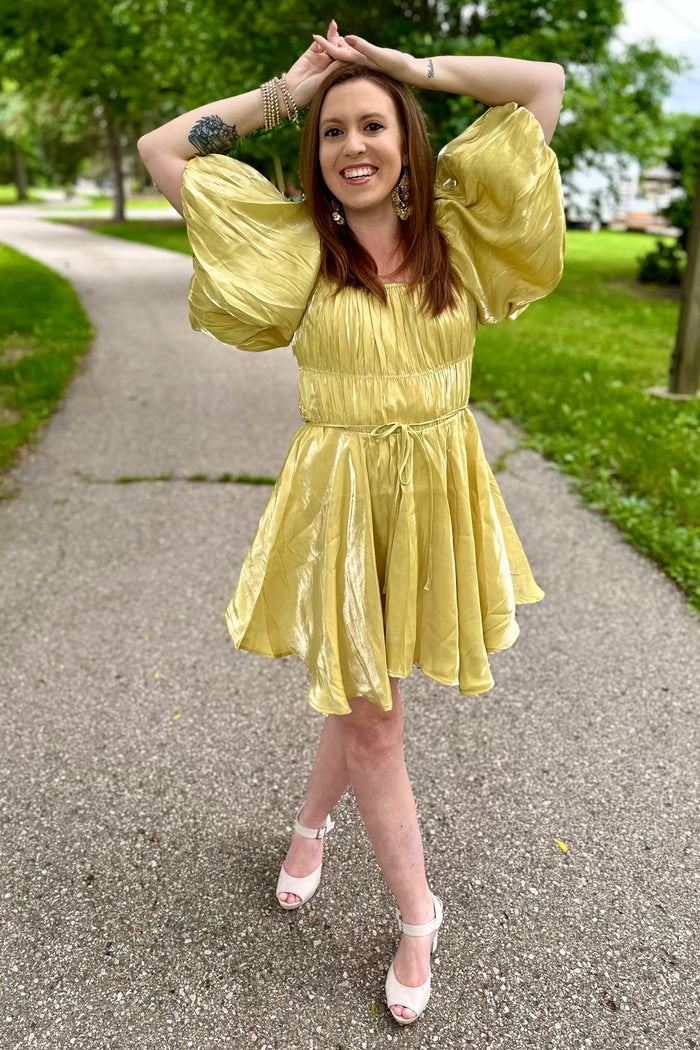 woman wearing a shiny light yellow short dress with puff sleeves and a cinched waist, posing outdoors on a path